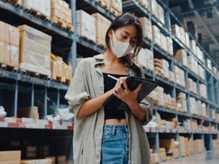 Young Asia businesswoman manager wear face mask looking for goods using digital tablet checking inventory levels stand in retail shopping center. Distribution, Logistics, Packages ready for shipment.
